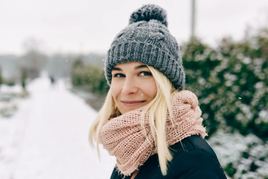 Close-up portrait of blonde beautiful young woman smiling wearing winter jacket, gray knitted hat and pink scarf, posing outdoors in the park on snowing winter day.