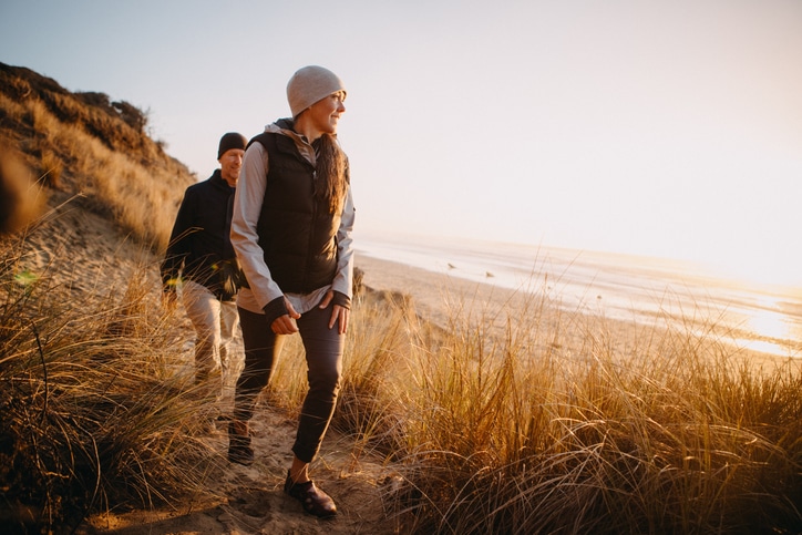 A woman wearing outdoorsy clothes and a vest and beanie travels along a beach with a man following behind her while looking at the ocean